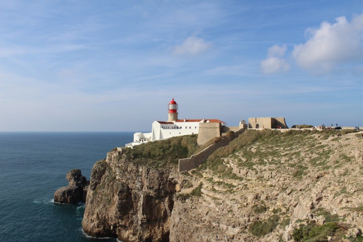 Lighthouse on rocky cliff overlooking ocean under blue sky with fluffy clouds.