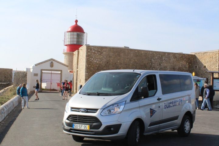 Van parked on road near a stone building with a red lighthouse and people walking.