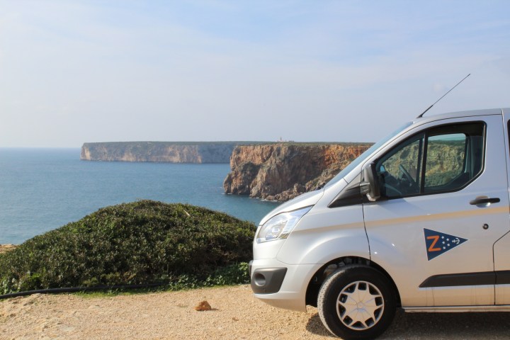 Van parked on cliffside overlooking ocean and distant cliffs under a clear sky.