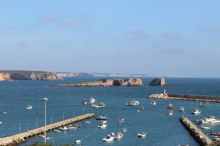 Coastal scene with boats in a harbor, rocky islands, and a lighthouse on a sunny day.