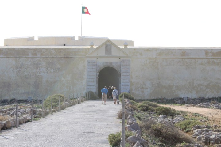 People walking towards an old fort with a flag on top, surrounded by a rocky path.