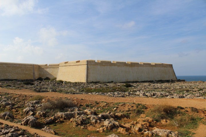 A historic stone fortress wall on a rocky landscape by the sea under a partly cloudy sky.