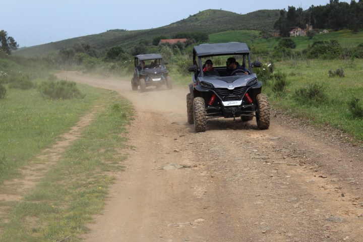 Two off-road vehicles drive on a dirt path with green hills in the background.
