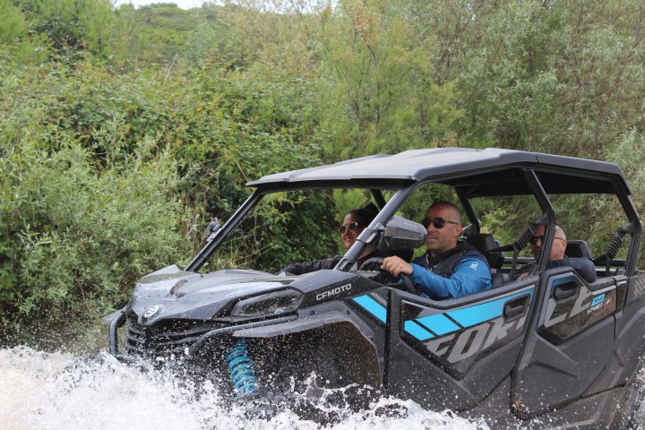 ATV with passengers driving through a water stream in a forested area.
