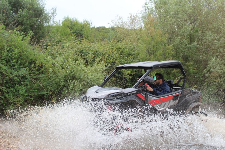 All-terrain vehicle splashes through water with driver on a forest trail.
